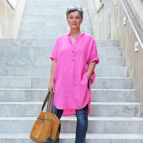 Frau trägt pinkfarbene Longbluse aus Leinen genäht nach schnittmuster oversize bluse von b-patterns. In der Hand hält sie eine große curryfarbene Tasche mit Reißverschluss. Dazu trägt sie schmale Jeans.